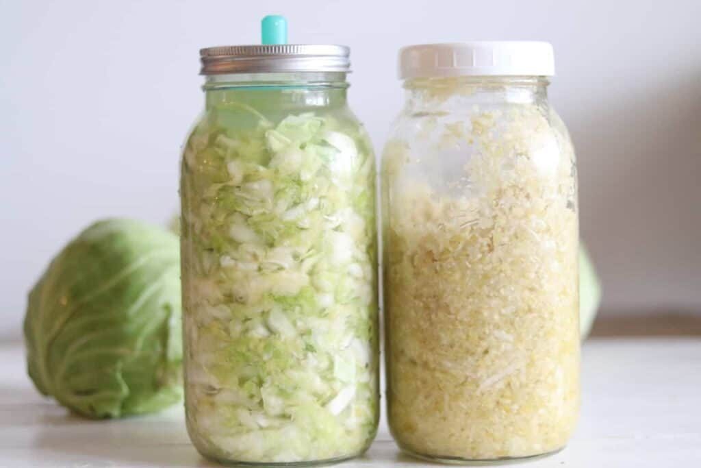 Two jars of sauerkraut sitting on white table with cabbage in background.