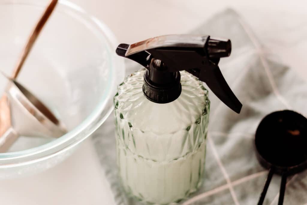 A green glass spray bottle next to soap scum remover supplies.