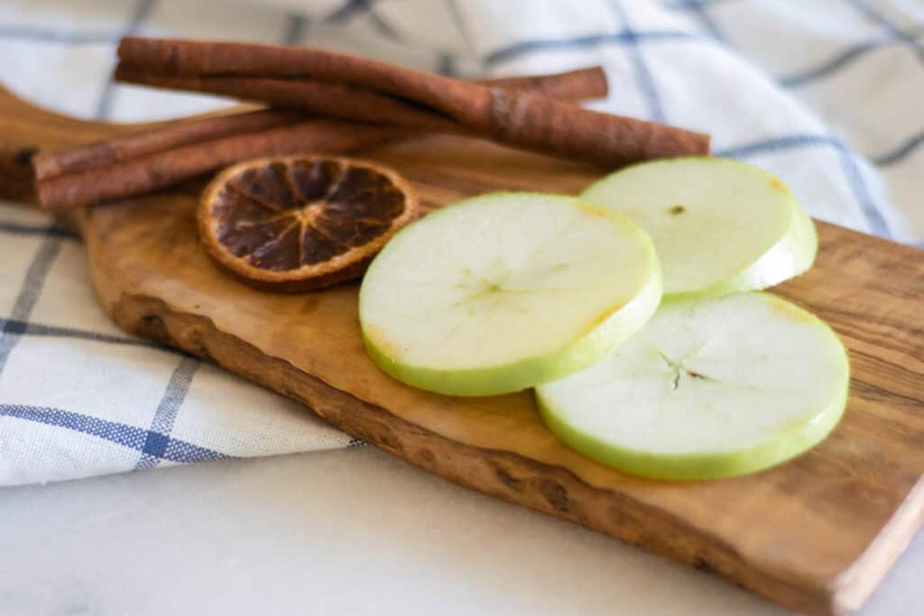 Sliced apples, oranges, and cinnamon sticks on a cutting board.