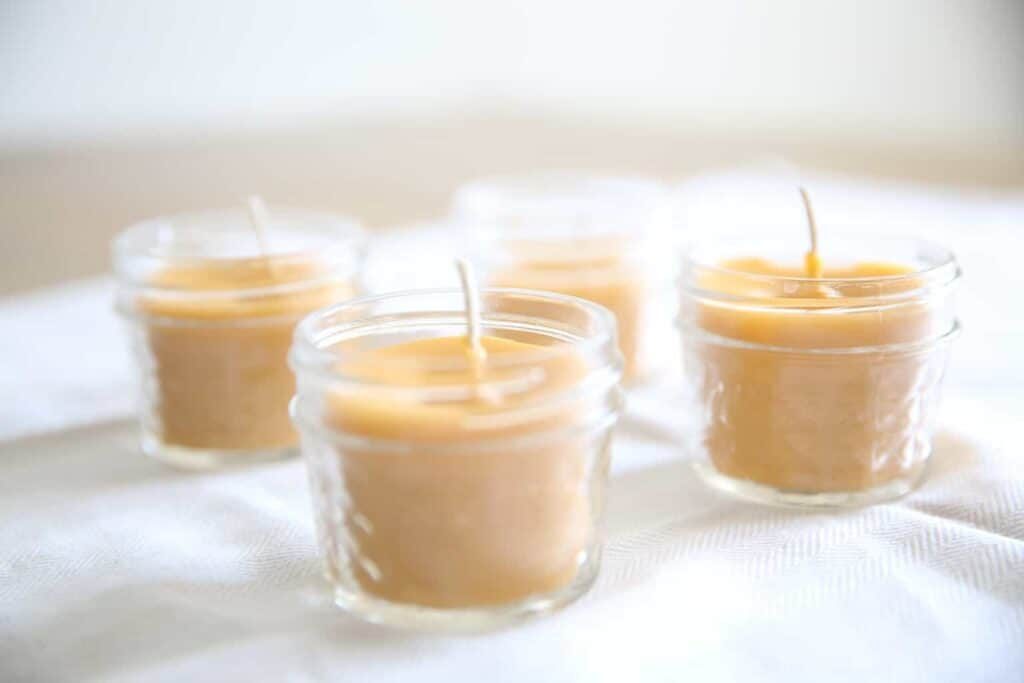 4 beeswax candles lined up on a white table in clear glass jars.