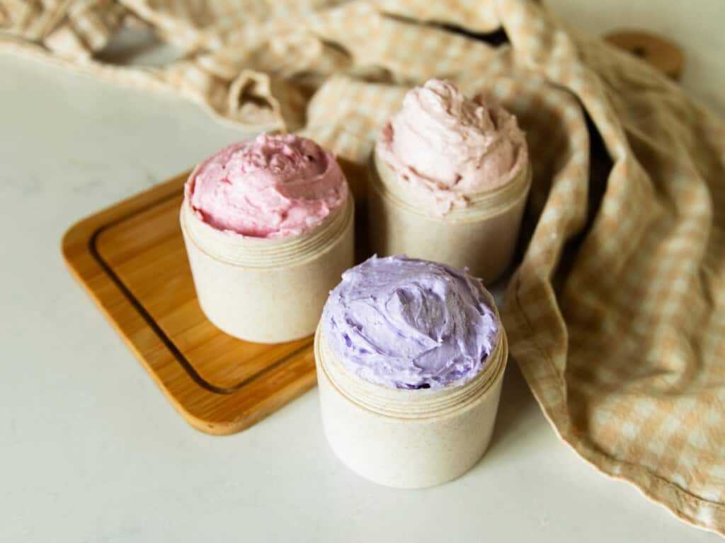 Three jars of different-colored whipped sugar soap on a wooden cutting board with a towel.
