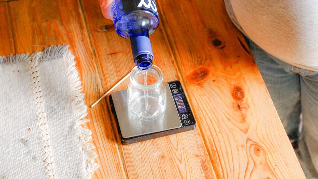 Vodka is poured into a glass jar sitting on a scale. The scale and jar sit on a wooden table with a linen table runner.