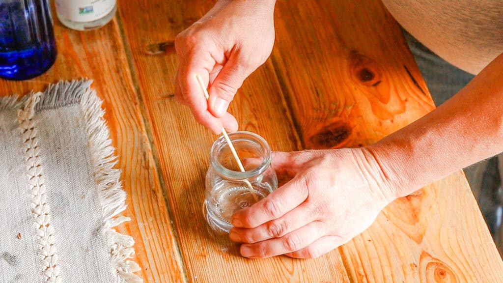 A hand is mixing ingredients in a glass jar for a homemade perfume recipe. The glass jar sits on a wooden table with a linen table runner.